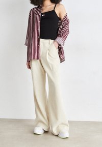 Canotta nera, camicia aperta a righe bordeaux, pantaloni larghi testurizzati color crema e scarpe bianche con dettagli verdi. Outfit casual e stratificato.