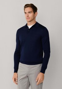 Granatowa, długosługa polo z kołnierzykiem, guziczkami i żebrowanym dołem; noszona na białej koszulce; gładka tekstura i dopasowany krój.