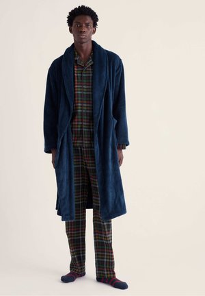 Homme portant un peignoir en peluche bleu foncé sur un pyjama à carreaux verts et rouges et des chaussettes rayées bleu marine et rouges, debout devant un fond uni.