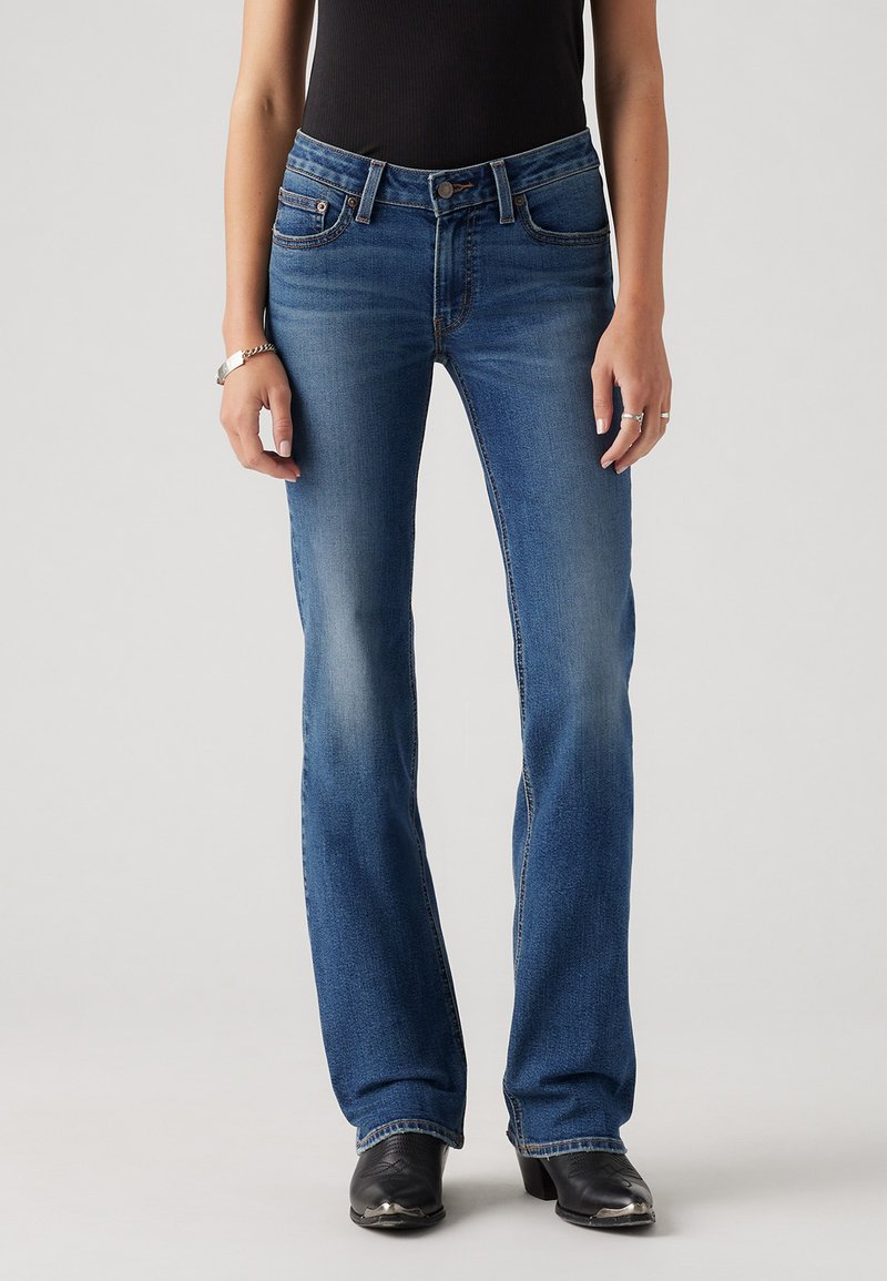Jean en denim bleu foncé ajusté avec une jambe évasée, taille mi-haute, cinq poches et coutures visibles. Porté avec des bottines noires.