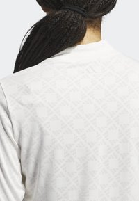 Bílá sportovní bunda s jemným geometrickým vzorem, hladkou texturou a vyraženým logem na límci.