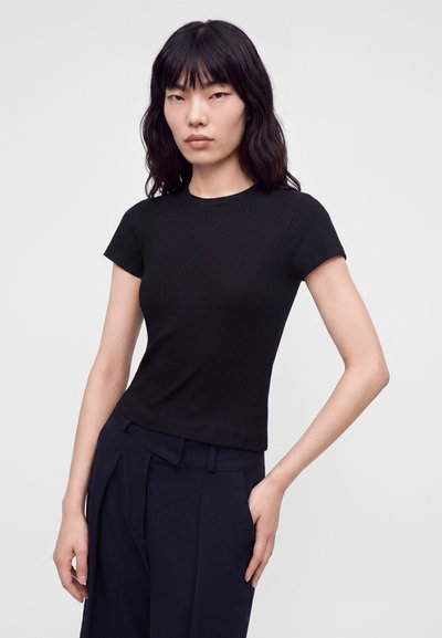 Femme aux cheveux noirs ondulés portant un haut noir ajusté à manches courtes et un pantalon sombre taille haute, debout avec une main dans la poche.