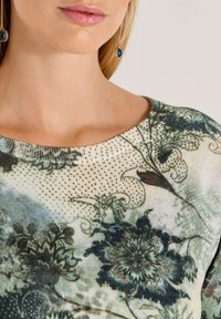 Vrouw in een bloeiende top met een gematigd groene en beige kleurstelling, met een close-up van haar nek, lippen en een paar blauwe steen oorknopen.
