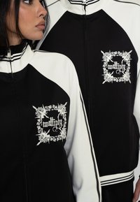 Sort-hvid zip-jakke med raglanærmer, stribet krave og detaljeret hvid broderi af ordet "multiply" på brystet.