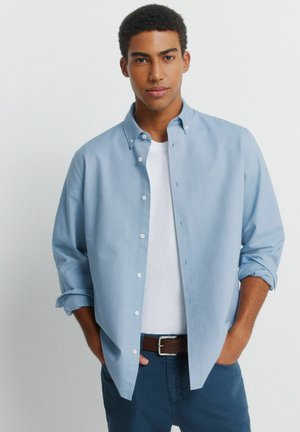 Junger Mann mit kurzen lockigen Haaren trägt ein hellblaues Hemd über einem weißen T-Shirt, die Hände in den Taschen, steht vor einem schlichten Hintergrund.