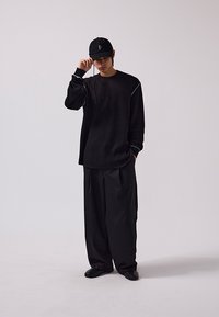 Schwarzes, übergroßes Langarmshirt aus strukturiertem Stoff mit kontrastierenden Nähten, kombiniert mit weiten schwarzen Hosen und einer schwarzen Mütze.