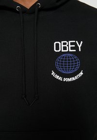 Černá mikina s kapucí s nápisem "OBEY" bílým písmem, obsahující modrý design globu a frázi "GLOBAL DOMINATION" bílým písmem pod tím. Šňůrky jsou černé.