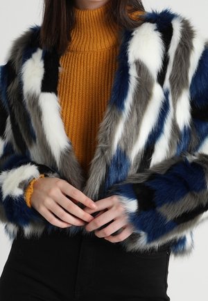 Personne portant une veste en fausse fourrure multicolore avec des rayures bleues, blanches, grises et noires sur un pull à col roulé côtelé jaune moutarde.