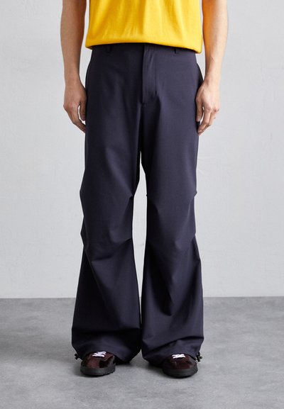 Wide-leg trousers in dark navy, made of smooth fabric. Features a classic fit, subtle front pleats, and paired with black and brown shoes.
