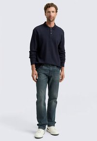 Tengerészkék hosszú ujjú póló háromgombos plackettel, elhalványult kék farmerrel és fehér tornacipővel párosítva. Laza szabás és puha anyag.