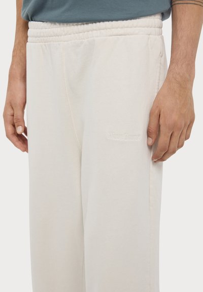 Light beige sweatpants with a textured finish, elastic waistband, and subtle logo detail near the pocket. Shape is loose-fitting.