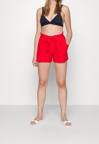 Femme portant un short rouge taille haute avec une ceinture nouée, un bralette noir et des sandales blanches à enfiler, se tenant contre un fond blanc uni.