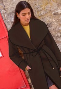 Palton maro cu o guler mare, detalii negre și o curea. Purtat peste un pulover galben, cu o textură netedă și bine definită.