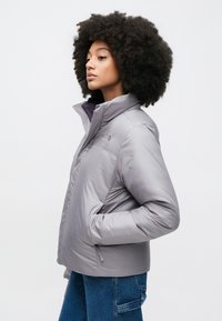 Γκρι μπουφάν με φουσκωτό ύφασμα, ψηλό γιακά, κλείσιμο με φερμουάρ και πλαϊνές τσέπες. Διαθέτει λογότυπο στο πάνω αριστερό μέρος του στήθους και έχει λείο, γυαλιστερό υλικό.