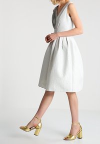 Robe blanche texturée avec un décolleté en V profond et des détails plissés ; associée à des sandales à talons blocs dorés brillants.