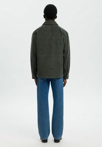 Persona di spalle che indossa una giacca camicia in corduroy verde scuro, jeans a gamba dritta blu e scarpe nere su uno sfondo bianco uniforme.