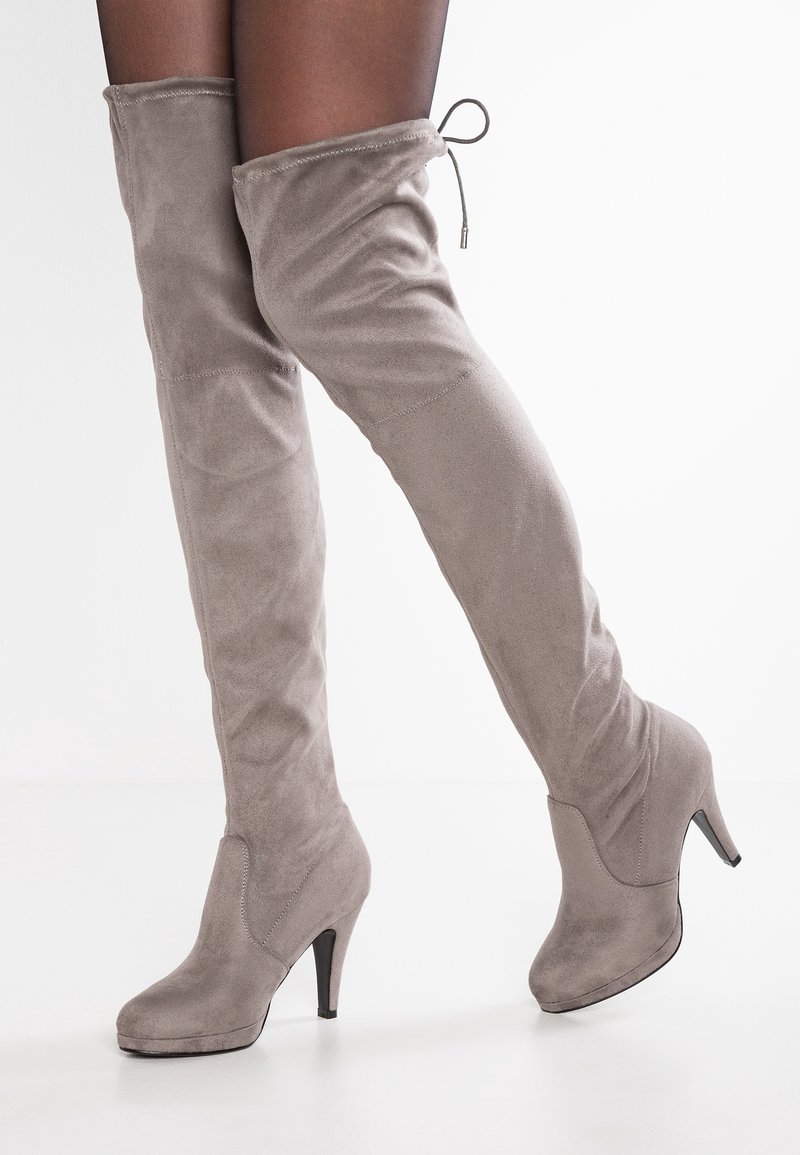 Nogi w przezroczystych czarnych rajstopach, noszące taupe zamszowe botki za kolano na wysokim obcasie z wiązaniem na sznurki na górze, na białym tle.