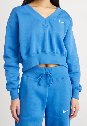Žena nosící modrou mikinu Nike s výstřihem do V a krátkým střihem a odpovídající tepláky se stahovací šňůrkou, která odhaluje břicho.