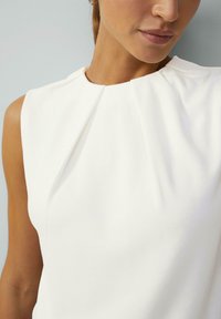 White sleeveless top featuring a pleated neckline, smooth fabric texture, and clean lines. Focus on simplicity and minimalistic design.