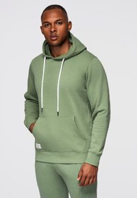 Homme portant un sweat à capuche vert sauge avec des cordons blancs et un pantalon de survêtement assorti, debout avec une main dans la poche devant un fond uni.
