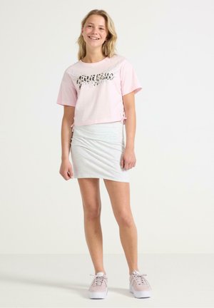 Rosa kortärmad t-shirt med ett tryckt mönster, kombinerad med en ljusgrå, samlad minikjol. Modellen bär beige sneakers.