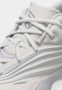 Primer plano de una zapatilla deportiva blanca con suela texturizada, cordones cruzados y un logo geométrico plateado en la lengüeta acolchada.