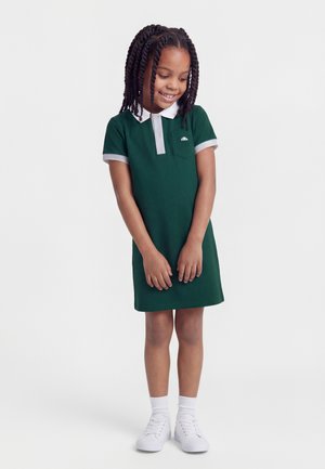 Ragazza giovane con capelli intrecciati che indossa un vestito polo verde con colletto e polsini bianchi, in piedi e sorridente su sfondo semplice.