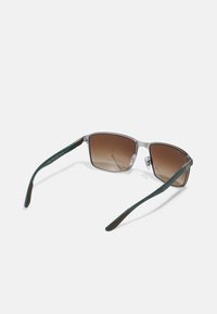 Ochelari de soare cu ramă metalică, lentile pătrate, nuanță gradient de maro, brațe verzi și accente din cauciuc maron la vârfuri.