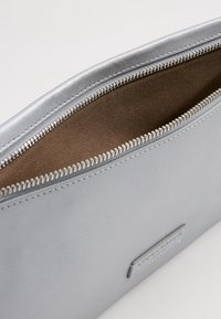 Silverfärgad konstläderpåse med brunt tygfoder, med dragkedjestängning och en liten präglad logotypdetalj på ena sidan.