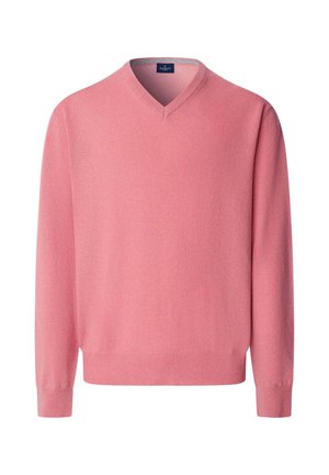 Hombre con suéter rosa de cuello en V, camisa a rayas azules, pantalones verde oscuro y zapatos marrones, de pie frente a una pared gris lisa.