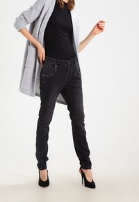 Světle šedý cardigan přes černý top, doplněný slim-fit černými džíny s kovovými detaily. Celý outfit dotváří černé lodičky na vysokém podpatku.