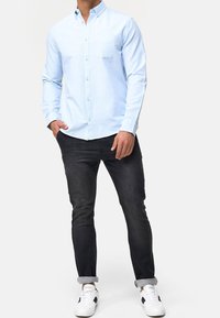 Helles blaues Hemd mit Knöpfen und einer Brusttasche, kombiniert mit dunklen Slim-Fit-Jeans und weißen Sneakers mit schwarzen Akzenten. Glatte Textur.