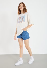 Oversized cream t-shirt with a palm tree graphic, paired with blue denim shorts and white sneakers with scalloped socks. Simple design.