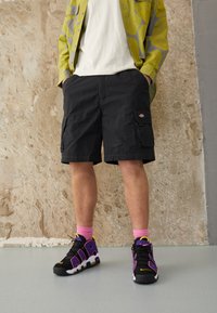 Black cargo shorts with side pockets, worn with black, pink, and yellow sneakers. The top features a patterned yellow jacket over a white shirt.