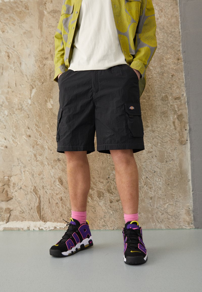 Black cargo shorts with side pockets, worn with black, pink, and yellow sneakers. The top features a patterned yellow jacket over a white shirt.