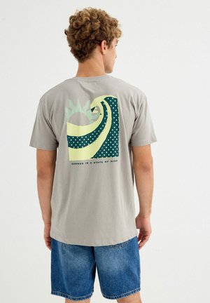 Joven con cabello rizado que lleva una camiseta gris con un gráfico estilizado de ola y sol y pantalones cortos vaqueros azules, de pie mirando hacia otro lado.