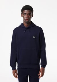 Sweat à capuche bleu marine en tissu doux, avec une poche kangourou, une capuche à cordon et un petit logo vert sur la poitrine. Design minimaliste.