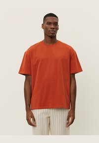 Tricou scurt cu maneci de culoare ruginită, din bumbac, cu decolteu rotund, croială confortabilă și detalii brodate discrete pe partea stângă a pieptului.