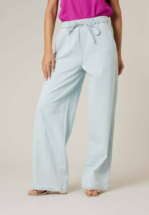 Femme portant un jean ample bleu clair à taille élastique avec cordon de serrage, associé à un haut rose et des sandales beiges.