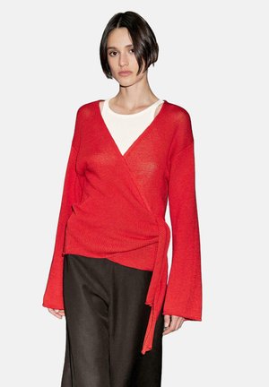Maglione avvolgente rosso con maniche lunghe, lavorazione a maglia testurizzata e scollatura a V profonda, indossato sopra una camicia bianca. Abbinato a pantaloni scuri.