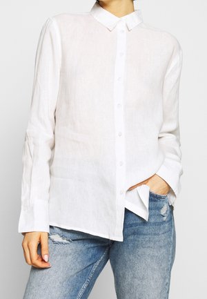 Person wearing a long-sleeve white button-up shirt and light blue ripped jeans, with one hand in pocket against neutral background.