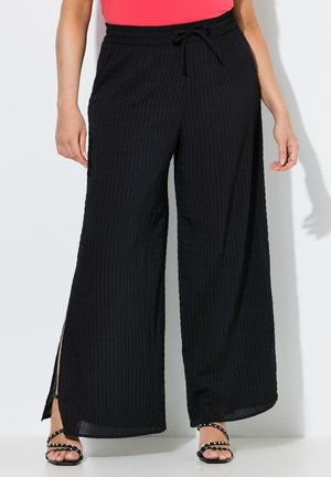 Vrouw die zwarte wijde broek met zijsplitten draagt, een roze top en zwarte open-toe sandalen met studs tegen een witte achtergrond.