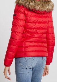 Rød puffjakke med hætte trimmet med pels, båret med blå denimjeans, som viser ryggen og armen med et lille logo patch på ærmet.