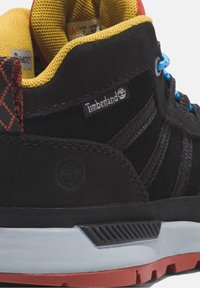 Czarny zamszowy sneaker z żółtą siateczkową wyściółką i niebieskimi sznurowadłami. Charakteryzuje się czerwonym akcentem na pięcie oraz logo Timberland. Podeszwa z czarnej, fakturowanej gumy.