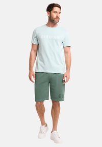 Helles blaues Baumwoll-T-Shirt mit weißem Logo, kombiniert mit grünen Baumwollshorts mit gedruckten Buchstaben und weißen Sneakern. Einfacher, lässiger Schnitt.