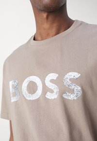 Muž v béžovém tričku s velkým, bílým, mírně opotřebovaným textem "BOSS" vytištěným na hrudi na jednobarevném pozadí.