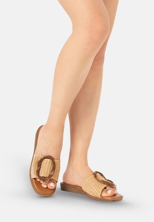 Mujer con sandalias tipo slide tejidas en color tostado con grandes hebillas circulares en tacones bajos marrones, de pie con las piernas cruzadas sobre un fondo blanco.