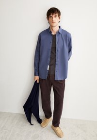 Camicia button-up azzurro chiaro sopra una maglietta noir, abbinata a pantaloni di velluto a coste marroni e scarpe slip-on color testa di moro, mentre tiene un maglione blu navy.
