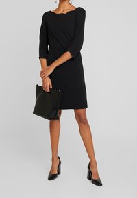 Černé šaty se scalloped výstřihem, tříčtvrtečními rukávy a přiléhavým siluetem. Doplněné černou kabelkou a podpatky.