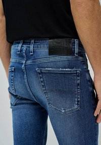 Jeans de denim azul ajustados con un desgaste mínimo, parche de cuero en la parte trasera etiquetado como "Salsa Jeans" y diseño estándar de bolsillos.